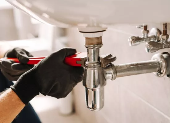 A plumber in black gloves provides plumbing services by fixing a sink pipe with a wrench in a bathroom